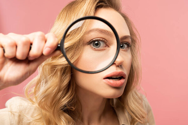 Blonde woman looking throughout magnifying glass isolated on pink
