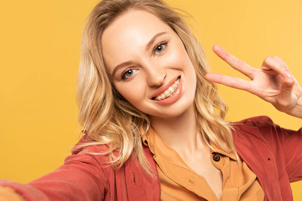 Smiling woman with dental braces showing peace sign while taking selfie isolated on yellow