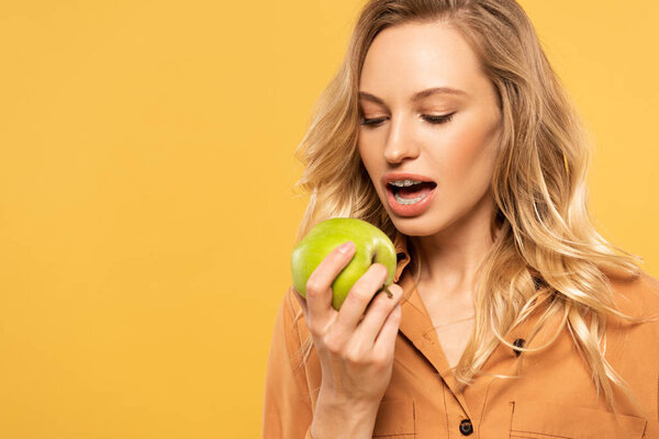Young woman with dental braces biting green apple isolated on yellow