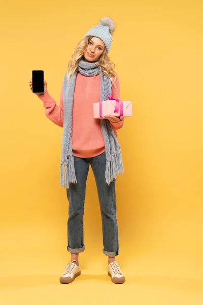 Blonde Woman Hat Scarf Holding Gift Box Smartphone Yellow Background — Stock Photo, Image