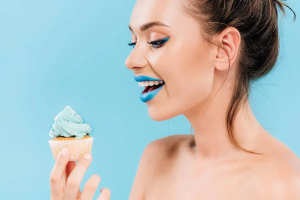 happy naked beautiful woman with blue lips looking at cupcake isolated on blue