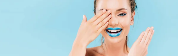 happy beautiful woman with blue lips covering eye with hand isolated on blue, panoramic shot