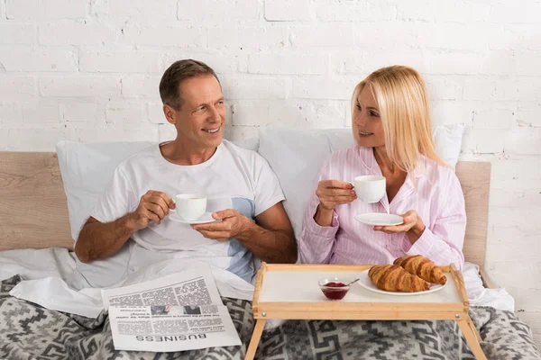 Gazeteyle Gülümseyen Olgun Çift Yatakta Kahvaltı Yapıyor — Stok fotoğraf