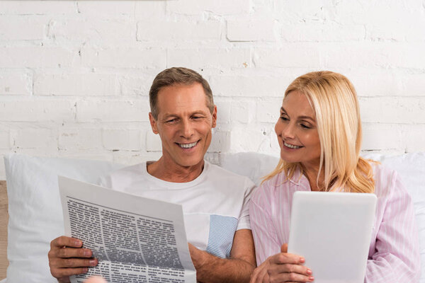 Man reading newspaper to wife with digital tablet on bed