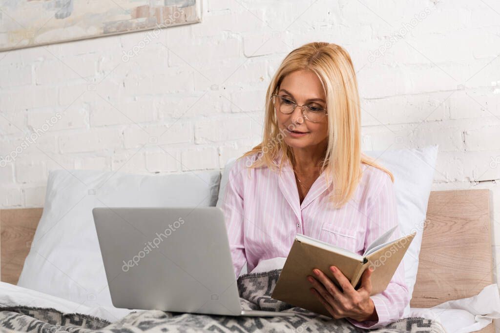 Woman in pajamas holding book and using laptop in bed