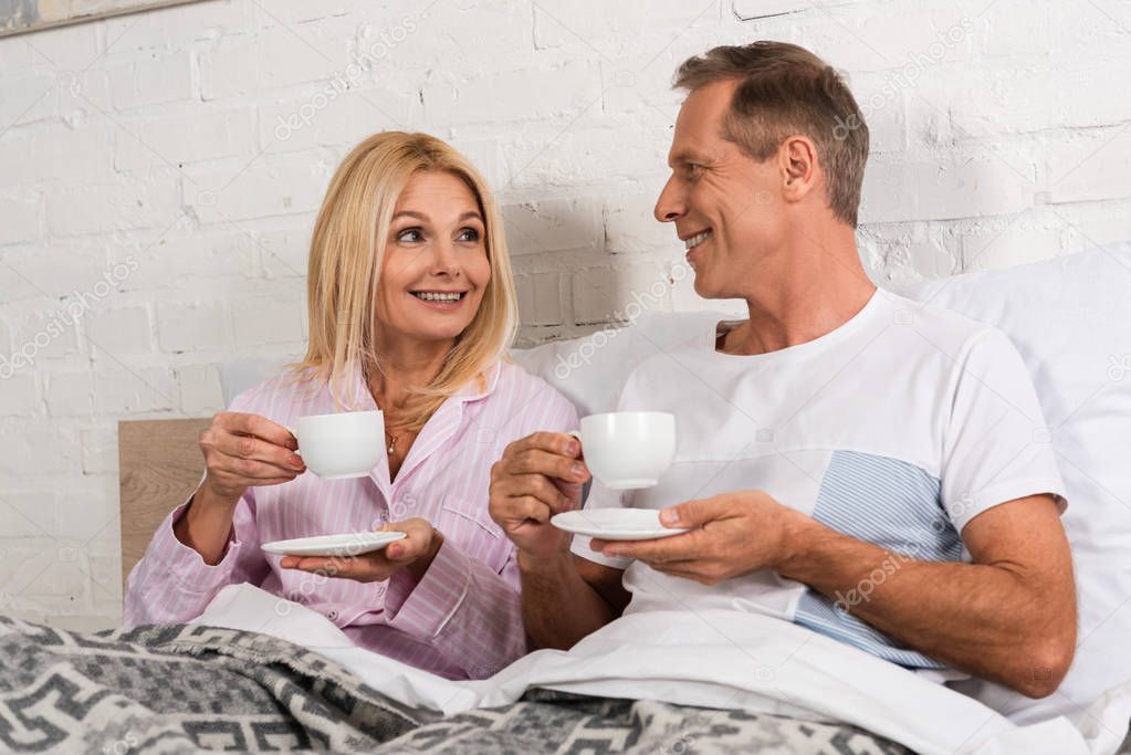 Smiling couple drinking coffee and looking at each other in bed