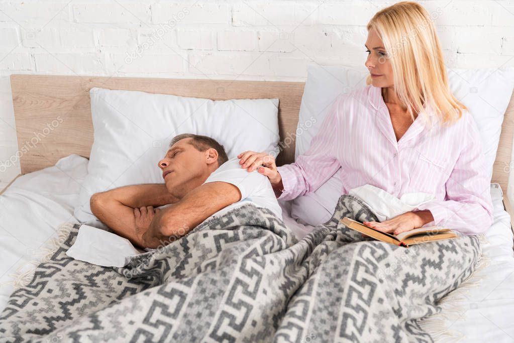 Woman with book waking up snoring husband in bed