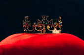 antique golden crown with gemstones on red pillow, isolated on black