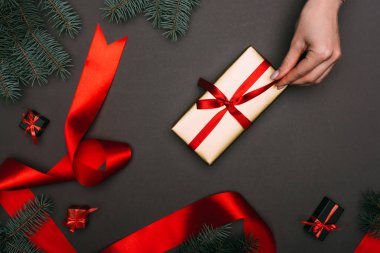 Kırpılmış bir manzara, siyah üzerine kırmızı kurdele ve ladin dallarıyla noel hediyeleri paketleyen bir kadın.