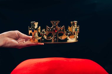 cropped view of woman holding golden crown with gemstones over red pillow, isolated on black clipart