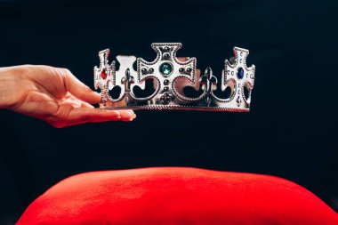 cropped view of woman holding silver crown with gemstones over red pillow, isolated on black clipart