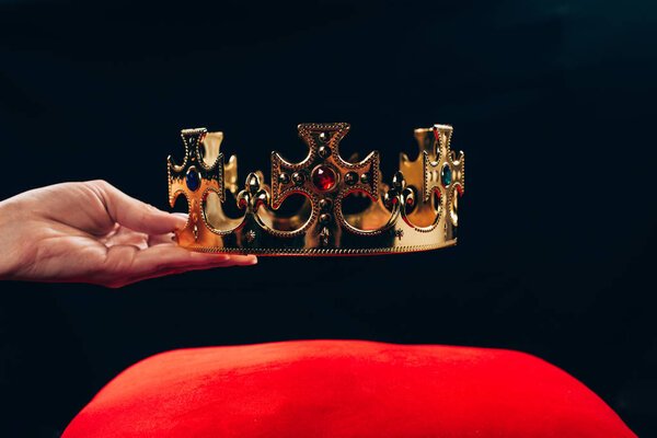 cropped view of woman holding golden crown with gemstones over red pillow, isolated on black