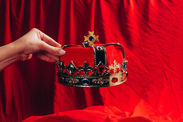 cropped view of woman holding golden crown with gemstones over red pillow 