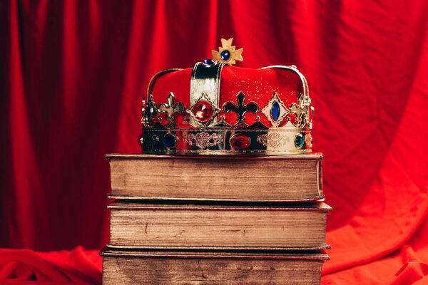 древняя золотая корона с драгоценными камнями на винтажных книгах на красной ткани
