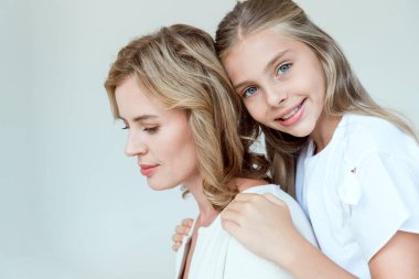 Gülümseyen kız annesine sarılıyor ve gri renkli kameraya bakıyor. 