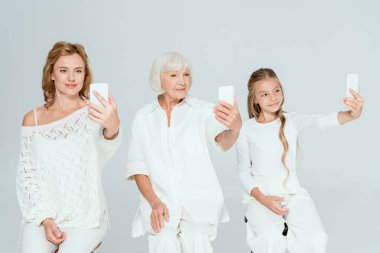 smiling granddaughter, mother and grandmother taking selfies isolated on grey  clipart