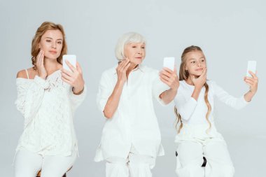 Gülümseyen torun, anne ve büyükanne selfie çekiyor ve gri üzerinde izole edilmiş yüzlere dokunuyor. 