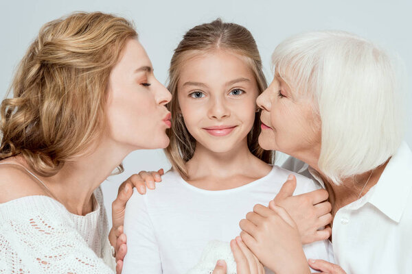 mother and grandmother kissing smiling granddaughter isolated on grey 