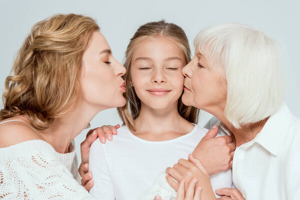 mother and grandmother kissing smiling granddaughter isolated on grey 