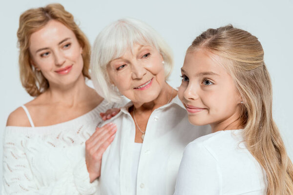 selective focus of smiling granddaughter, mother and grandmother looking at her on background isolated on grey 