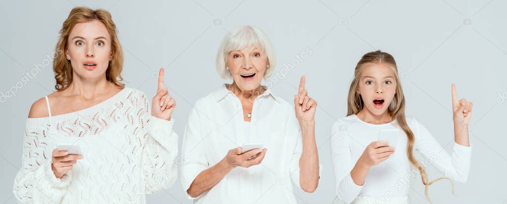panoramic shot of shocked granddaughter, mother and grandmother holding smartphones and showing idea gestures isolated on grey 