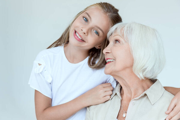 smiling granddaughter hugging grandmother and looking at camera isolated on grey 