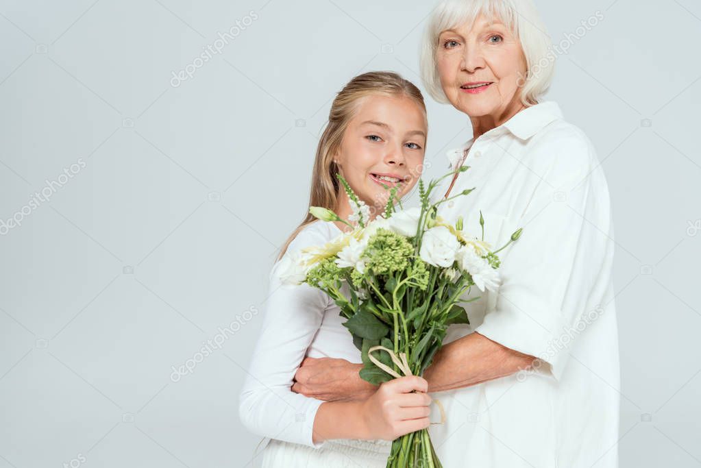 smiling grandmother hugging granddaughter with bouquet isolated on grey 