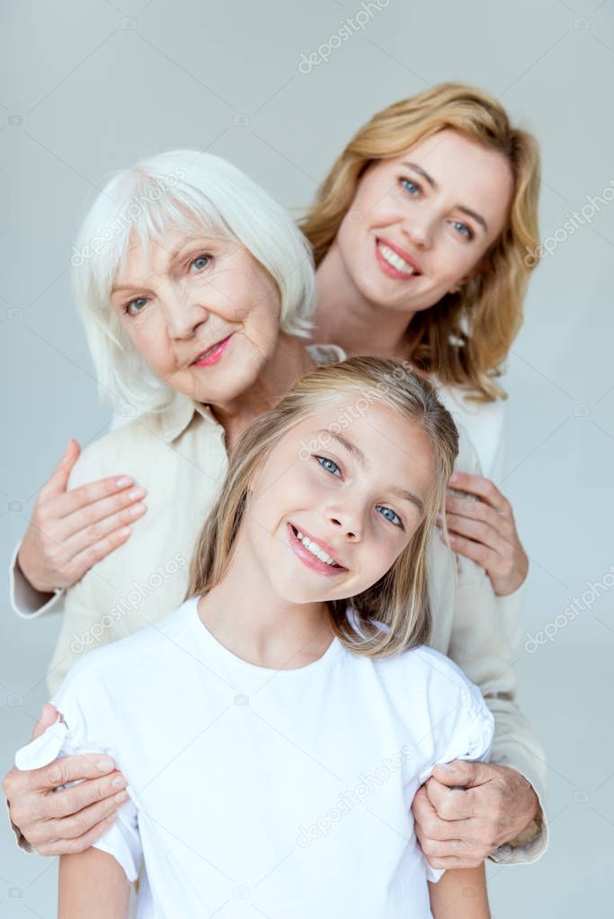 smiling granddaughter, grandmother and mother hugging isolated on grey 