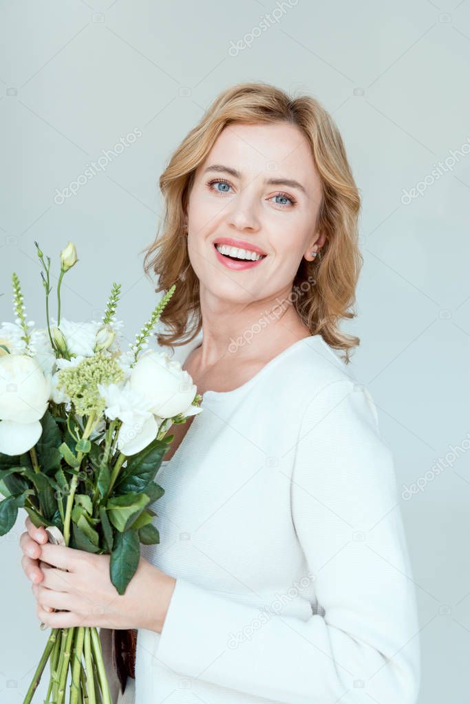 smiling and attractive woman with bouquet looking at camera isolated on grey