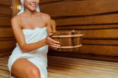 oříznutý pohled na usmívající se ženu v ručníku držící dřevěné umyvadlo v sauně 