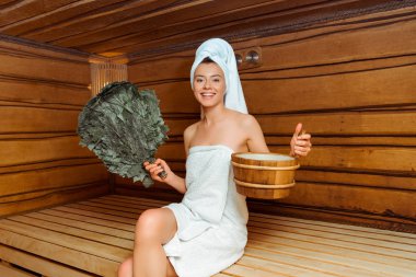 smiling woman in towels holding birch broom and washtub in sauna  clipart