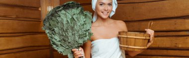 panoramic shot of smiling and attractive woman in towels holding washtub and birch broom in sauna  clipart