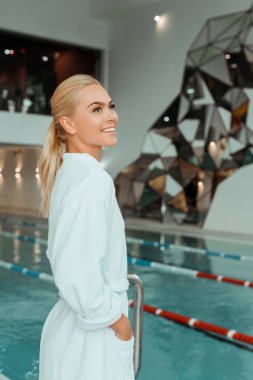 Beyaz bornozlu, çekici ve gülümseyen bir kadın kaplıcaya bakıyor. 