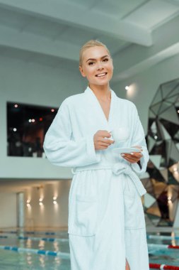 Beyaz bornozlu, kaplıcada kahve tutan çekici ve gülümseyen kadının düşük açılı görüntüsü. 