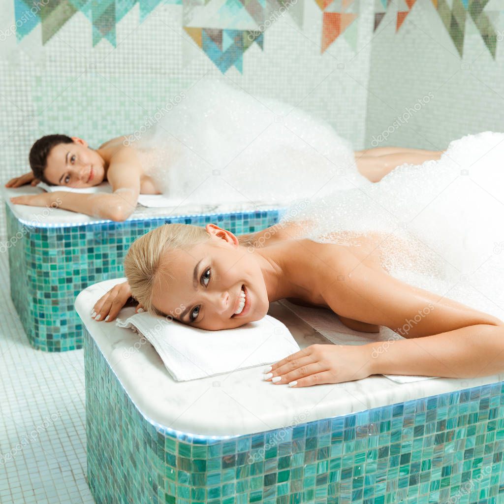 attractive and smiling friends lying with foam in Turkish bath