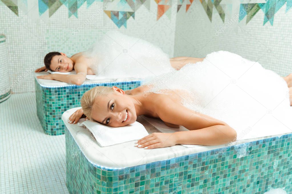 attractive and smiling friends lying with foam in Turkish bath
