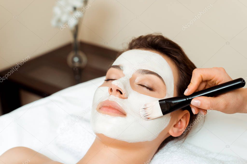 cropped view of cosmetologist applying face mask on attractive woman in spa 