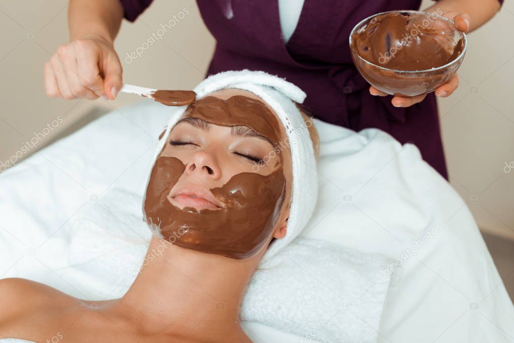cropped view of cosmetologist applying face mask on attractive woman in spa 