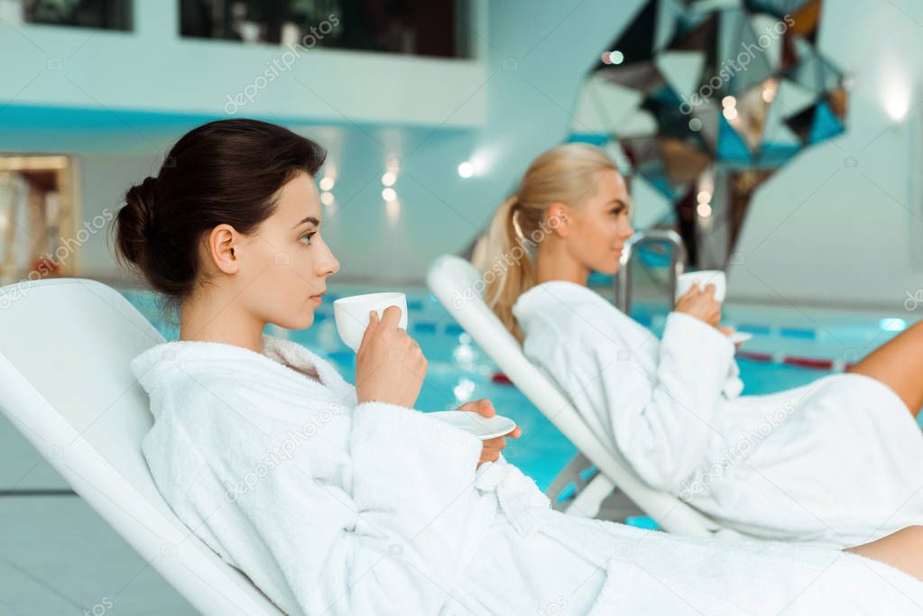 selective focus of attractive woman in white bathrobe holding cup and friend on background in spa 