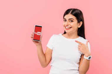 Kyiv, Ukrayna - 20 Eylül 2019: güzel esmer kız pembe üzerinde izole edilmiş uygulamayla akıllı telefonu işaret ediyor.