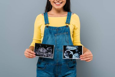 cropped view of smiling pregnant pretty girl holding fetal ultrasound images isolated on grey clipart