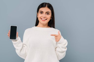 Beyaz kazaklı, gülümseyen güzel esmer kız gri ekranlı akıllı telefonu işaret ediyor.