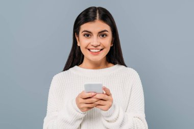Gülümseyen, beyaz kazaklı güzel esmer kız. Gri üzerine izole edilmiş akıllı telefon kullanıyor.
