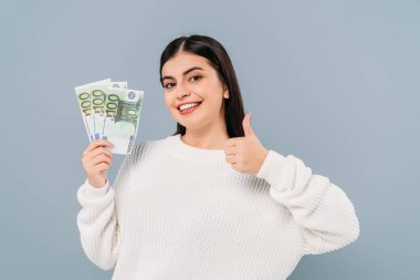 smiling pretty girl in white sweater holding euro banknotes and showing thumb up isolated on grey clipart
