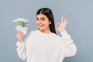 smiling pretty girl in white sweater holding euro banknotes and showing ok isolated on grey clipart