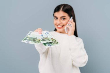 Seçici odak noktası, beyaz kazaklı güzel bir kızın euro banknotları vermesi ve gri üzerinde izole edilmiş akıllı telefondan konuşması.
