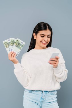 Elinde euro banknotları olan beyaz kazaklı güzel bir kız ve gri üzerinde izole edilmiş akıllı telefon kullanıyor.