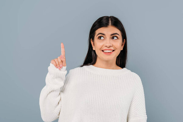 Улыбающаяся красивая брюнетка в белом свитере, указывающая пальцем вверх, изолированная на сером
