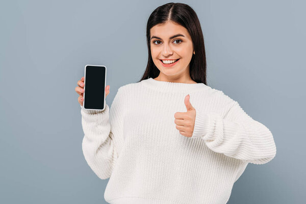 smiling pretty girl in white sweater showing smartphone with blank screen and thumb up isolated on grey