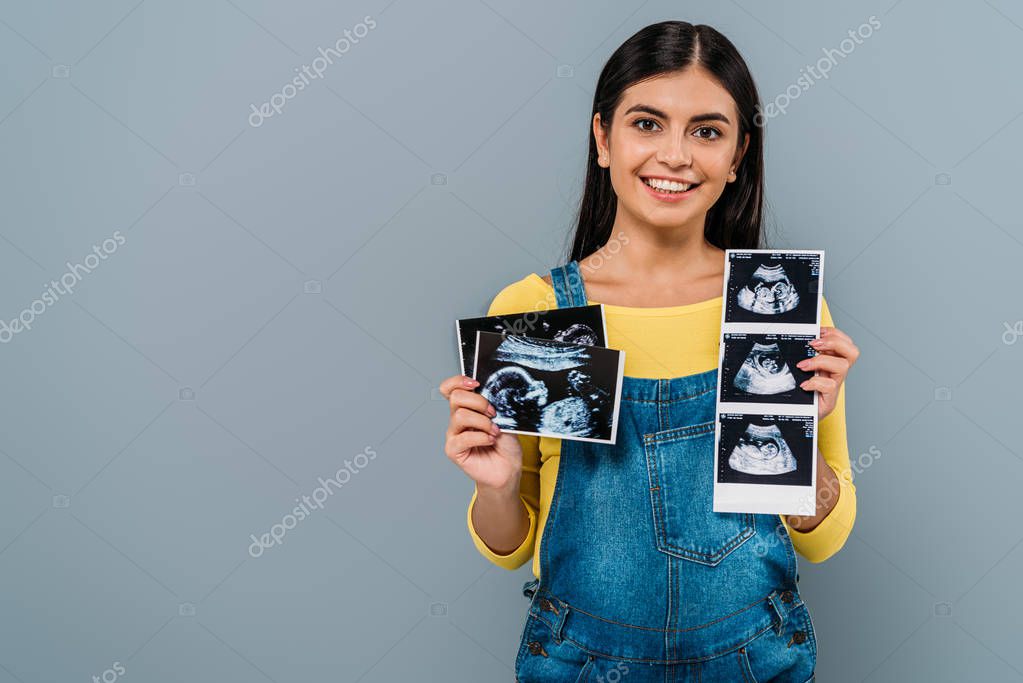 Smiling pregnant pretty girl holding fetal ultrasound images isolated on grey